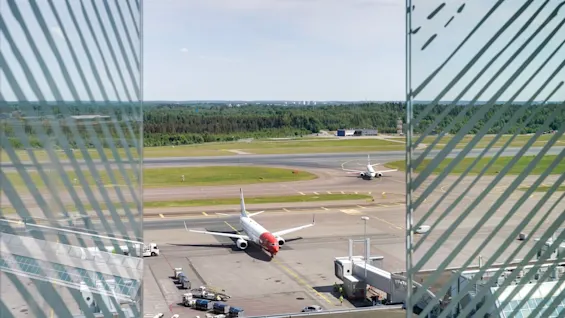Udsigt fra en lufthavnsterminal, der viser fly på landingsbanen og forpladsen, med en skov i baggrunden.