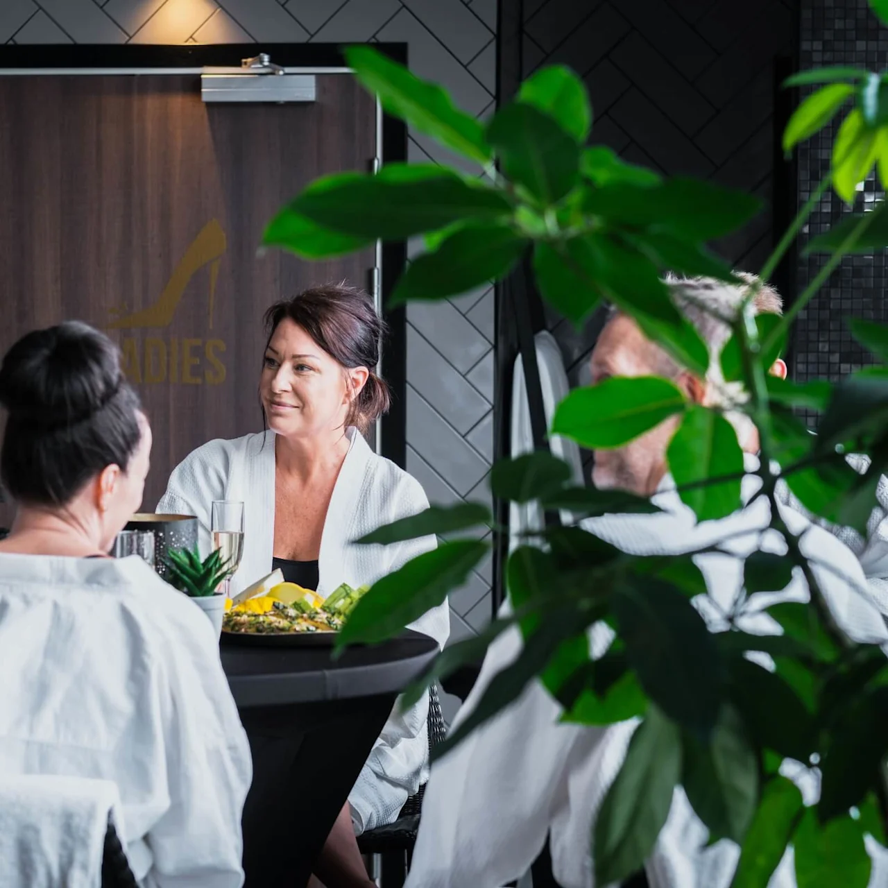 Four people in white robes sit at a table, conversing in a spa-like setting. A sign reading "LADIES" with a high-heeled shoe is visible on a door in the background.