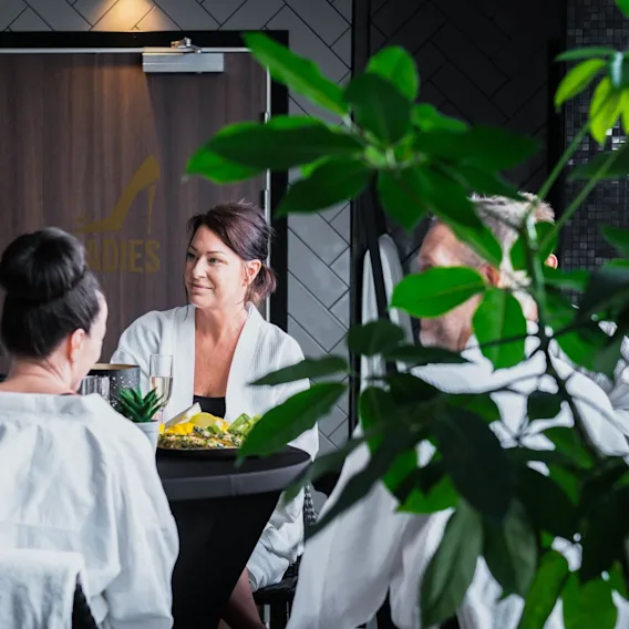 Fyra personer i vita morgonrockar sitter vid ett bord och samtalar i en spa-liknande miljö. En skylt med texten "LADIES" och en högklackad sko syns på en dörr i bakgrunden.