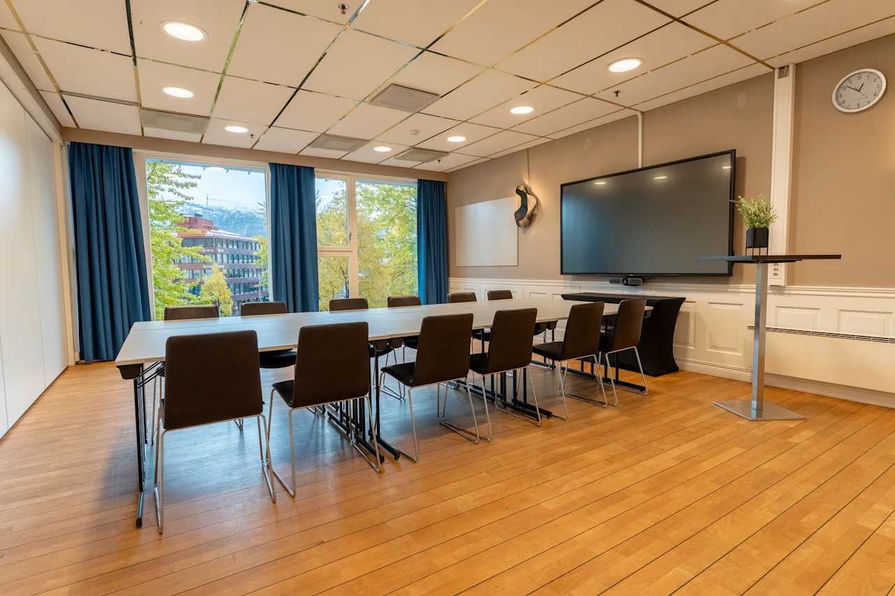 Moderni kokoushuone, jossa on pitkä pöytä, tuolit, suuri näyttö ja luonnonkaunis näkymä rakennuksiin ja vuorille ulkona.