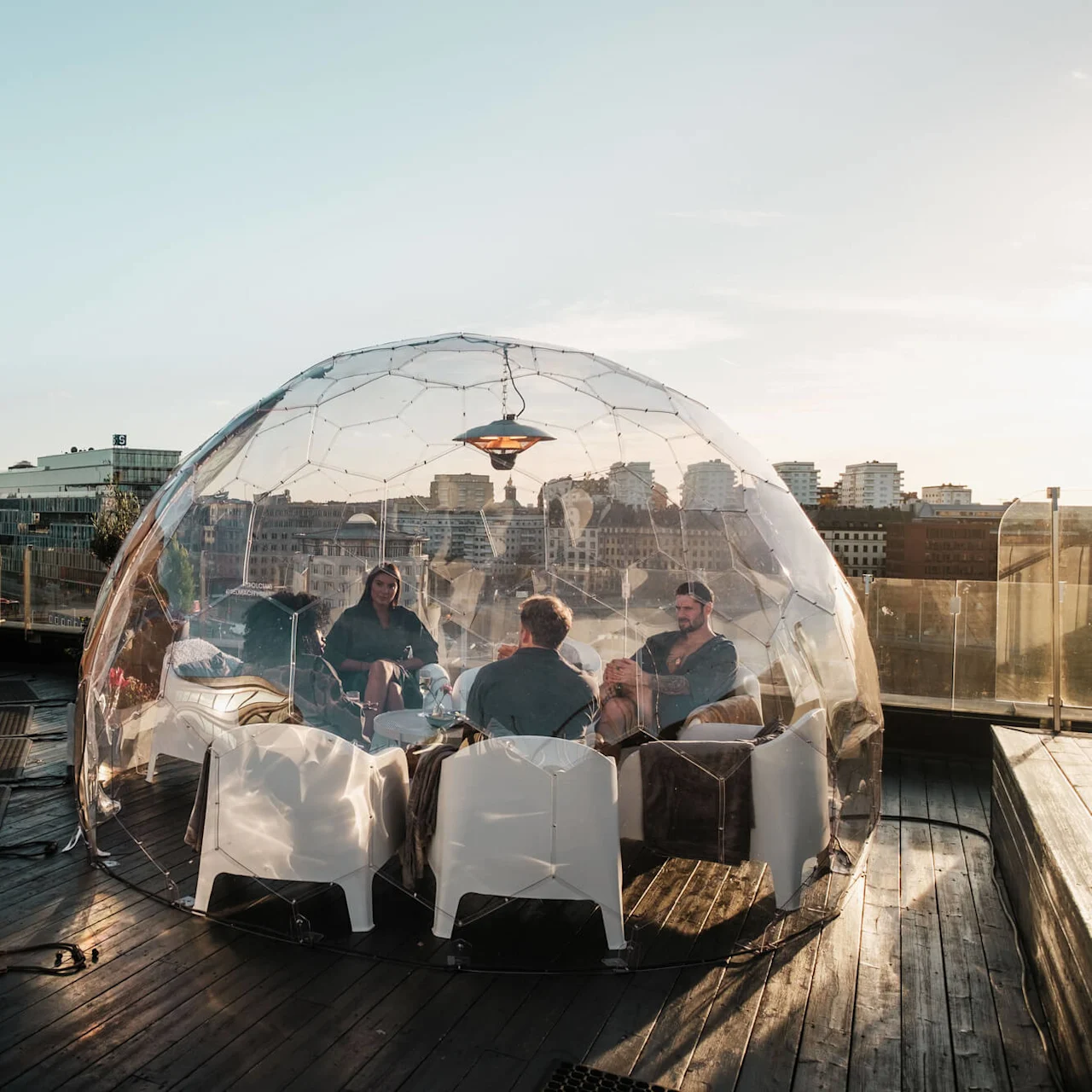 Transparent kupol skyddar fyra personer som sitter och pratar på en takterrass med utsikt över staden. En trästruktur och fler kupoler kompletterar utomhusmiljön.