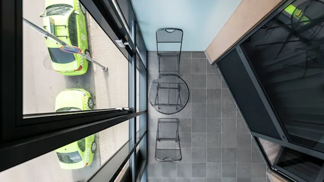 A black metal table and chairs set on a tiled balcony overlooking a street. Two bright green cars are parked below.