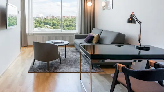 En stue med en grå sofa med fargerike puter og et rundt bord under et stort vindu som viser byens silhuett. En svart skrivebordslampe står på et stilfullt skrivebord.