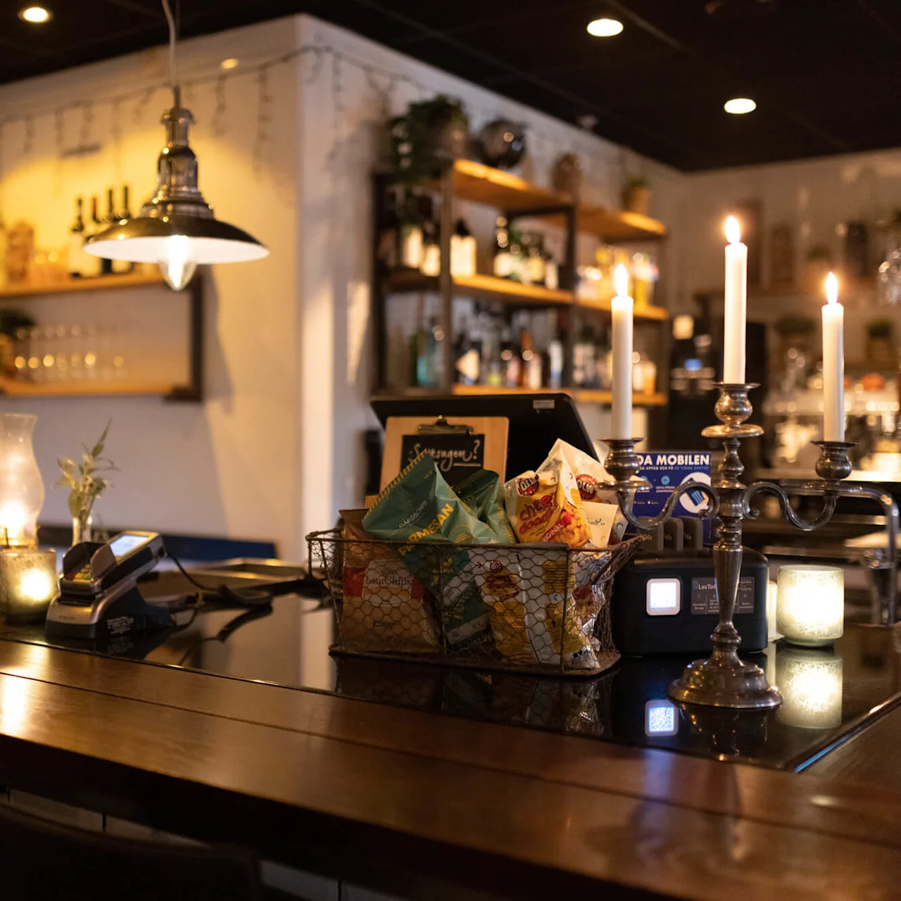 A cozy bar with lit candles and hanging lamps offers a warm atmosphere, complete with snacks and bar equipment.