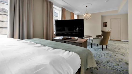 En säng med grönt överkast framför en stor TV i ett elegant inrett rum med gardiner, ljuskrona och fåtölj.