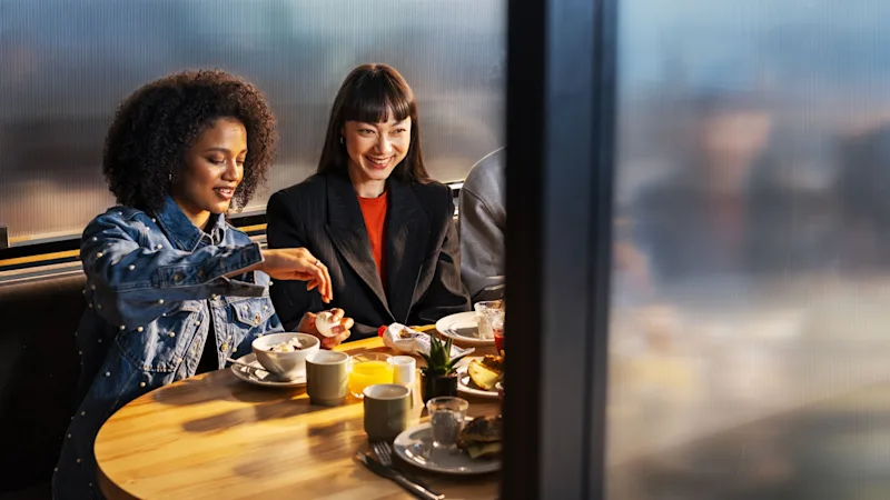 Två personer njuter av en måltid vid ett runt träbord med diverse frukostartiklar, i ett mjukt belyst matrum. En person tillsätter socker i sin dryck.