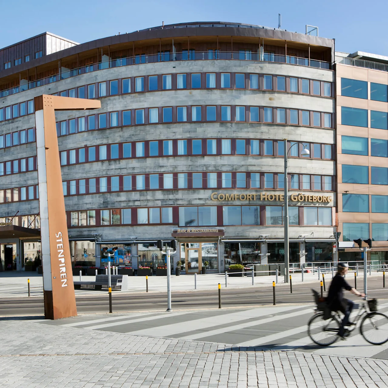 En cyklist kör förbi Comfort Hotel Göteborg, en vinklig byggnad med reflekterande fönster på en solig dag. I närheten finns en träskulptur märkt "STENPIREN".