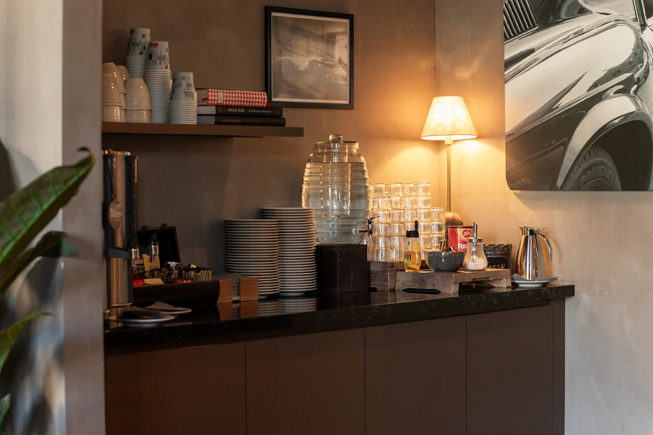 En indbydende selvbetjeningsstation med drikkevarer, kopper og tallerkener, oplyst af en varm lampe. Synlig tekst: SUGAR, TEA, COFFEE, N1.