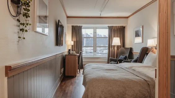 A cozy hotel room with a large bed, a desk, and chairs, featuring a window view of a snowy mountain landscape.