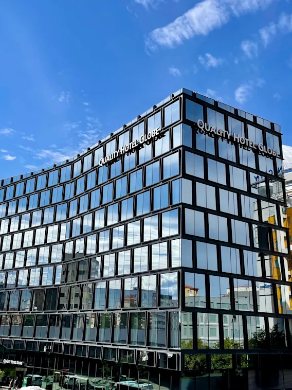 En moderne glassbygning viser "QUALITY HOTEL GLOBE" på fasaden, som reflekterer solfylt blå himmel. Bygningen står mot en bybakgrunn med tilstøtende gule og hvite strukturer.