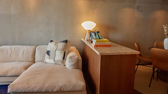 En beige sektionssoffa med en mönstrad kudde står mot en texturerad vägg, bredvid en träbänk med en lysande lampa och böcker märkta "OSTERIA 16" och "EATALY" i ett mysigt vardagsrum.