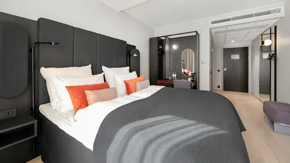 A neatly made bed with gray and orange pillows sits in a modern hotel room. A mirror, desk, and door are in the background.