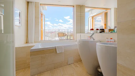 Ett lugnt badrum med ett stort fönster bredvid badkaret, som erbjuder en ljus utsikt över en balkong och stad under en blå himmel.