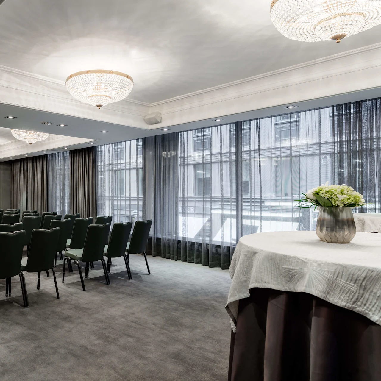 Elegant meeting room with rows of green chairs facing a projector screen, crystal chandeliers, and large windows with sheer curtains.