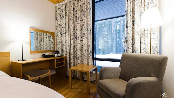 Gray armchair sits next to a wooden table in a cozy room. Curtains with a nature pattern cover large windows, and a desk with a chair and lamp is nearby.