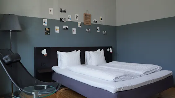 A neatly made double bed in a modern bedroom with pillows aligned, surrounded by a dark chair and round glass table. Wall displays are decorated with small art pieces.