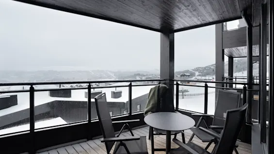 En træterrasse med to stole og et lille rundt bord står under et tag. En frakke hænger over en stol. Sneklædte bygninger og bakker er synlige udenfor.