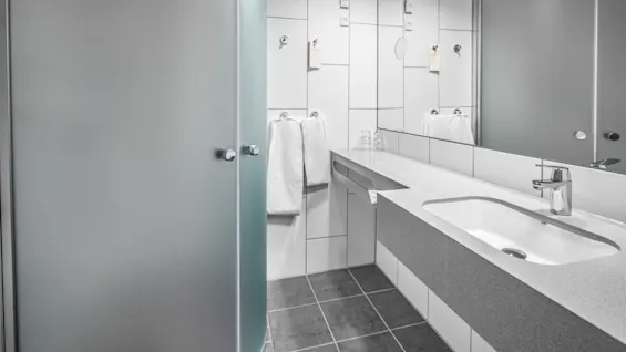Modern bathroom featuring a frosted glass shower, a countertop with an integrated sink, and mirrored walls. White towels and a dark tiled floor complete the sleek design.