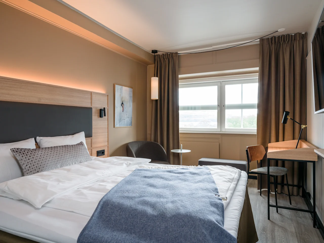 En säng är prydligt bäddad med vit och blå sängkläder, placerad i ett modernt hotellrum. Rummet har ett skrivbord med en stol, ett fönster med gardiner och väggkonst.