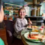 Barn smiler, mens de holder en pølse. De spiser med en anden person på en farverig, livlig restaurant. Omkransede glas med juice, tallerkener med mad og moderne indretning fylder omgivelserne.