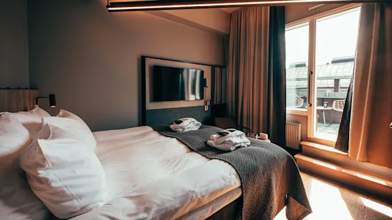 En ryddig seng med et teksturert teppe ligger i et koselig hotellrom. I nærheten er en flatskjerm-TV montert på veggen, og en solfylt balkong er synlig gjennom glassdører.