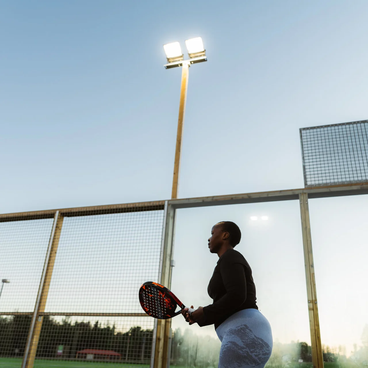 En person som holder en padelracket står klar til å spille på en utendørsbane med høye nettingvegger, under lyse stadionlys mot en klar himmel.