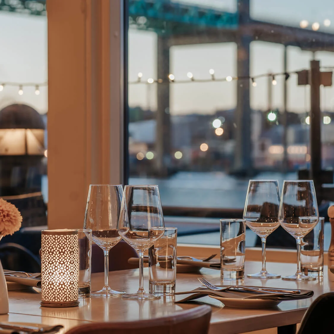 A set dining table with wine glasses and a lit candle overlooks a waterfront view and bridge at dusk, providing a serene ambiance.