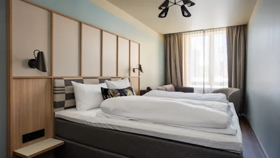 Ett mysigt hotellrum med en stor säng, en stilren sänggavel och ett fönster som släpper in naturligt ljus. Perfekt för avkoppling.