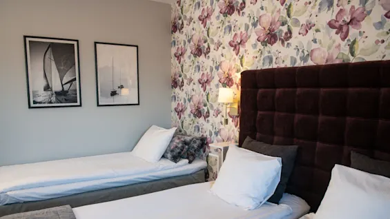 Two neatly made single beds stand in a hotel room with floral wallpaper, a padded headboard, and framed sailboat photographs. A wall lamp softly illuminates the space.
