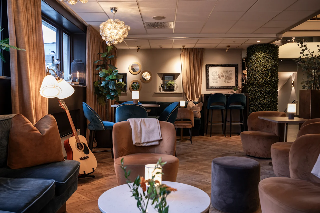 A warm and inviting lounge area at Home Hotel Grand Bodø, featuring comfortable seating, soft lighting, and a guitar, suggesting a relaxed atmosphere.