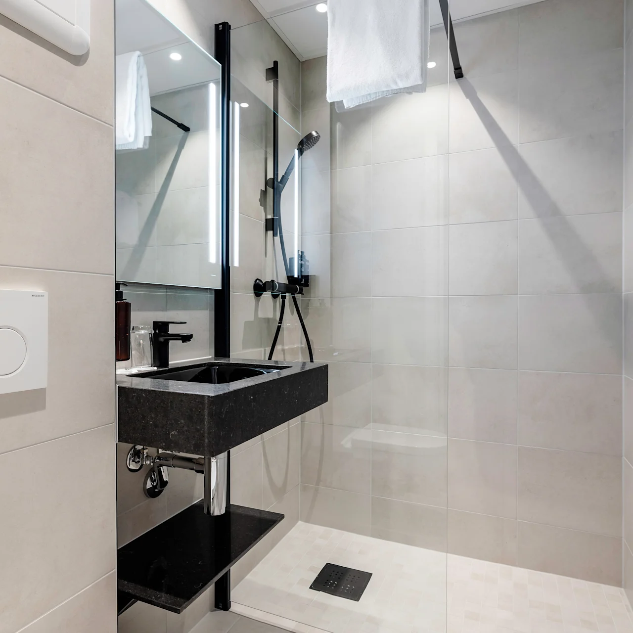 Modern bathroom features a black sink beside a glass-enclosed shower. Context includes beige tiled walls, a circular dual-flush toilet button, and neatly folded towels on the right wall.