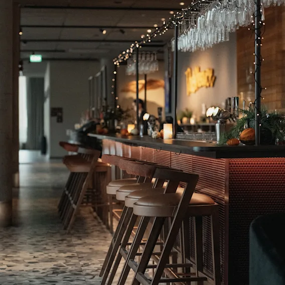En stilren hotellbar med barstolar i trä, hängande vinglas med ljusslinga och ett terrazzogolv.