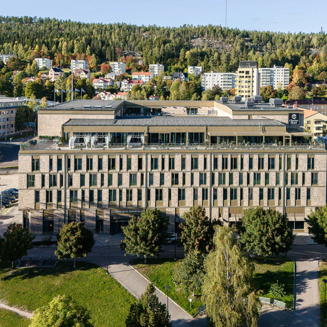 Et stort, rektangulært hotel står i et grønt forstadsområde med træer og lave bygninger. Synlig tekst: "CLARION HOTEL SUNDSVALL" og "e Clarion Hotel."