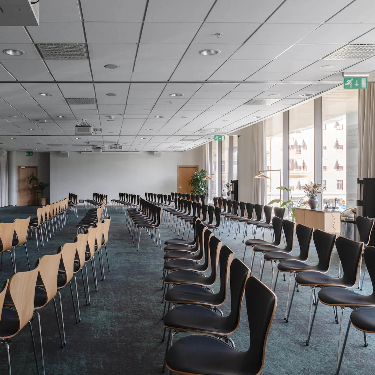 Rader av tomma stolar står mot en scen i ett konferensrum. Stora fönster släpper in naturligt ljus, och krukväxter dekorerar rummet.