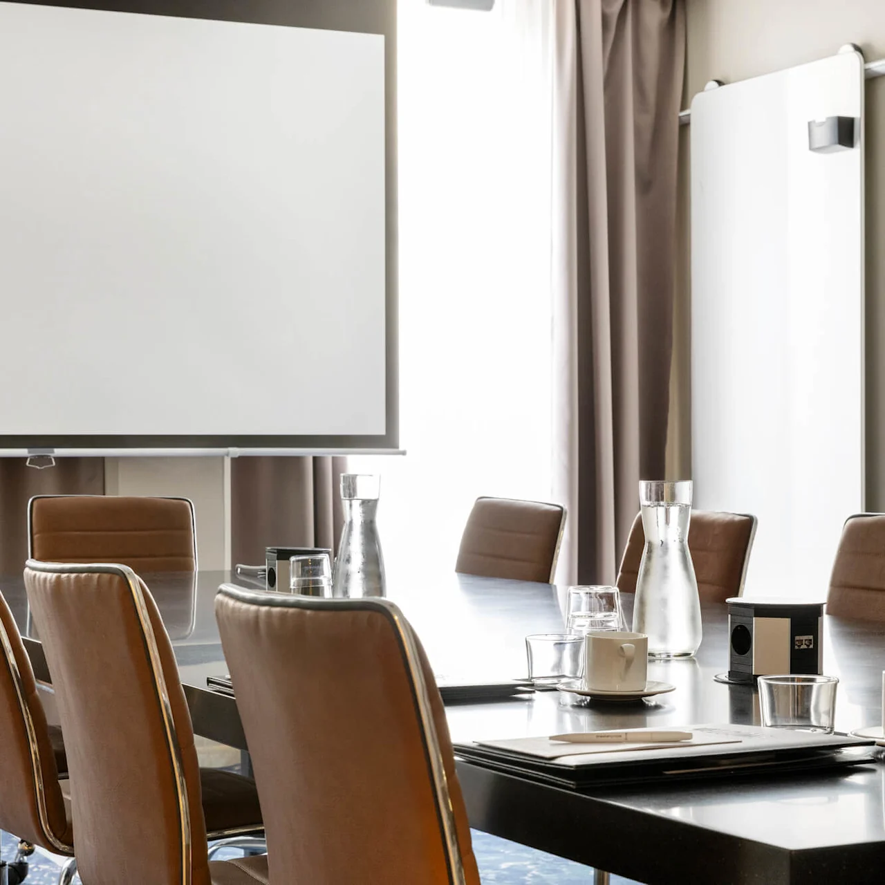 A conference room with brown chairs surrounding a large table, featuring water carafes and cups. A projection screen and whiteboard are in the background, with curtains partially drawn.