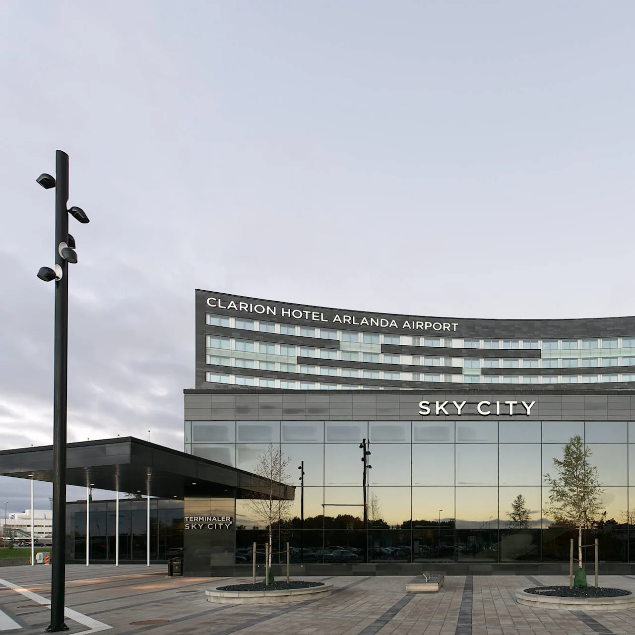 A modern hotel building stands with large glass windows. The sign reads "CLARION HOTEL ARLANDA AIRPORT" and "SKY CITY." Trees are planted nearby.