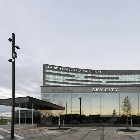 En modern hotelbyggnad står med stora glasfönster. Skylten lyder "CLARION HOTEL ARLANDA AIRPORT" och "SKY CITY." Träd är planterade i närheten.