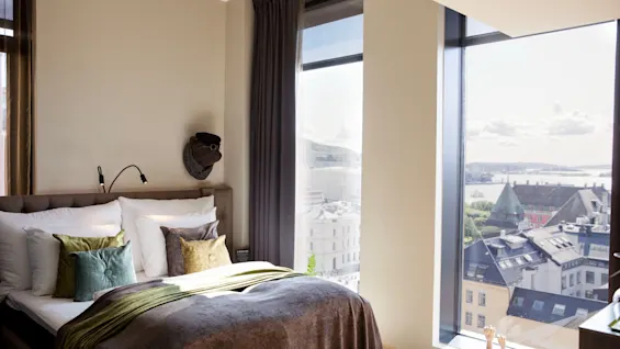 A neatly made bed with colorful pillows and a brown throw blanket stands by large windows. The windows offer a cityscape view of rooftops and a distant body of water under a clear sky.