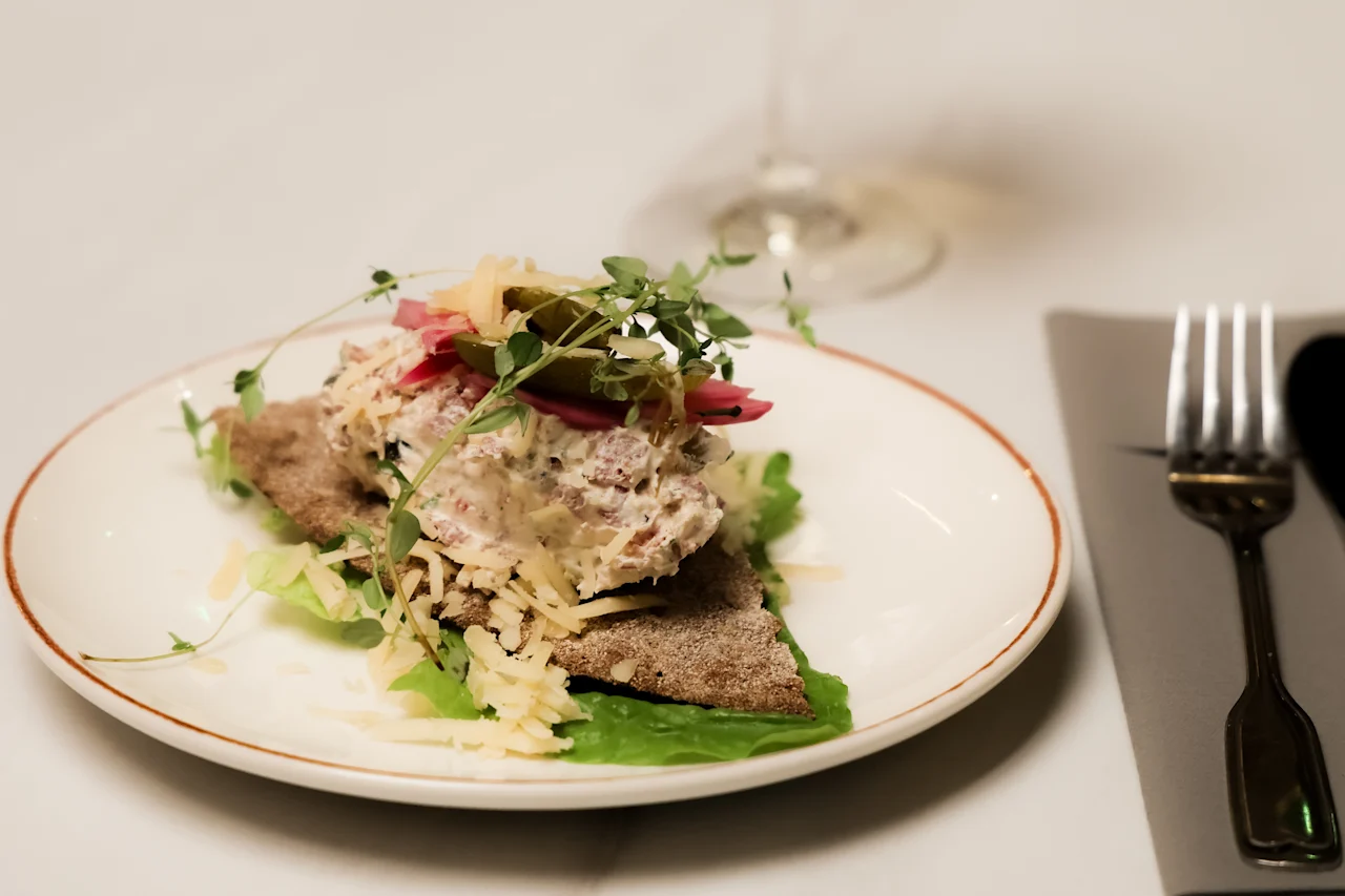 A dish of bread topped with creamy salad, pickled vegetables, microgreens, and grated cheese on a lettuce leaf. Positioned on a white plate beside a wine glass and cutlery.