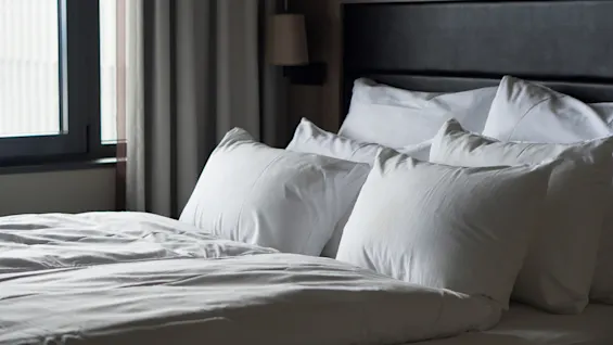A neatly made bed with white pillows and duvet against a wooden headboard, situated near a window with closed curtains in a softly lit bedroom.