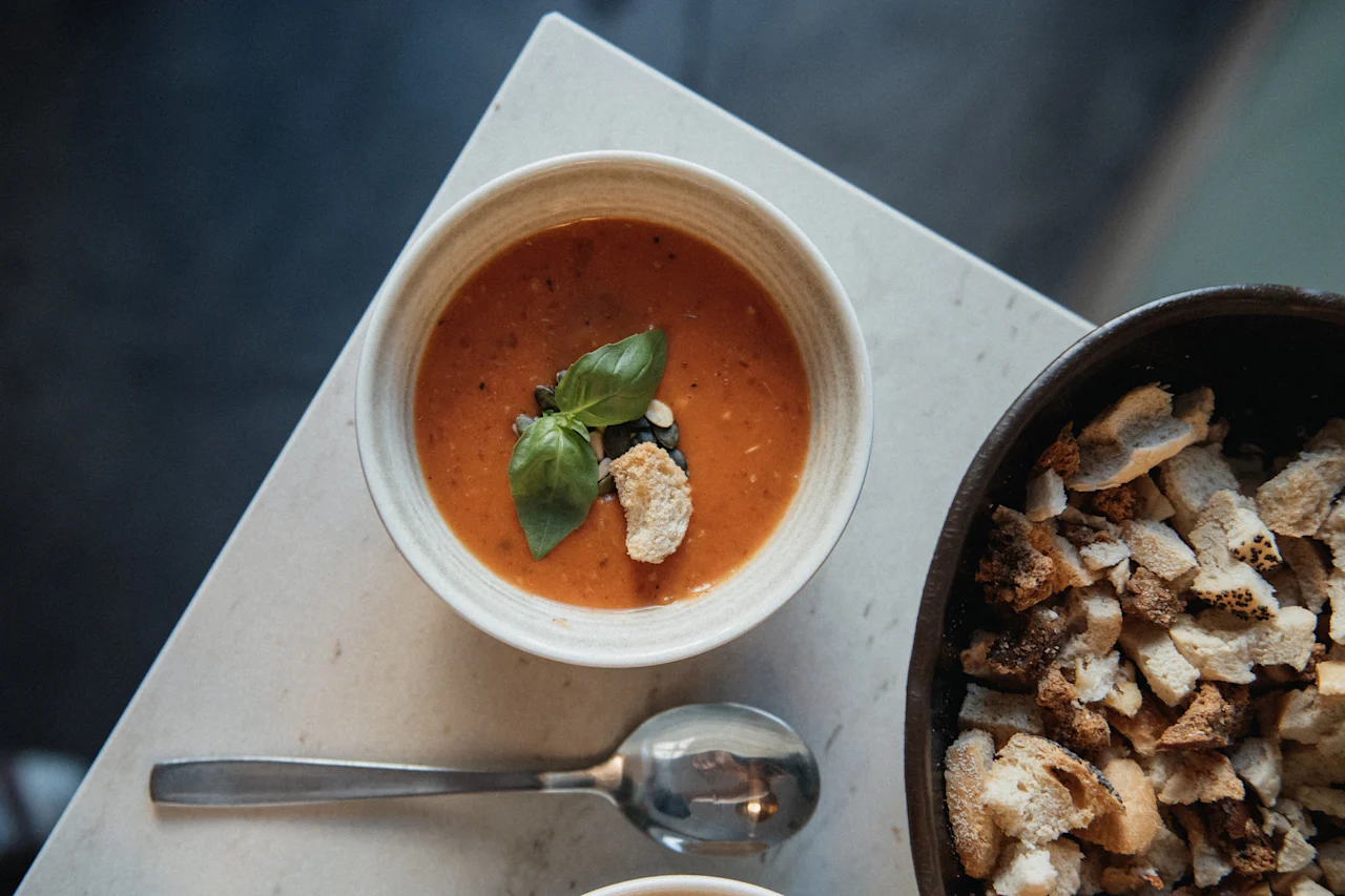 En skål tomatsoppa garnerad med basilika och krutonger, bredvid en skål med brödbitar. Home Hotel Post.