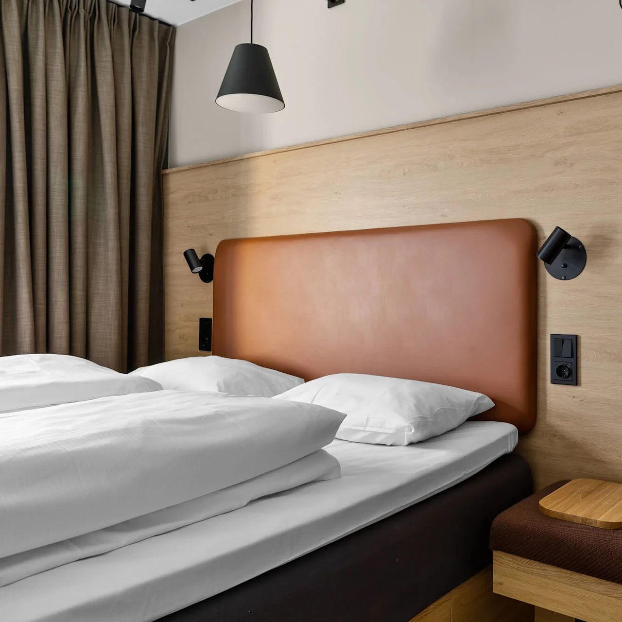 A bed with white linens and a brown headboard is flanked by black reading lamps. It's adjacent to brown curtains, a wooden wall panel, and a cushioned bench.