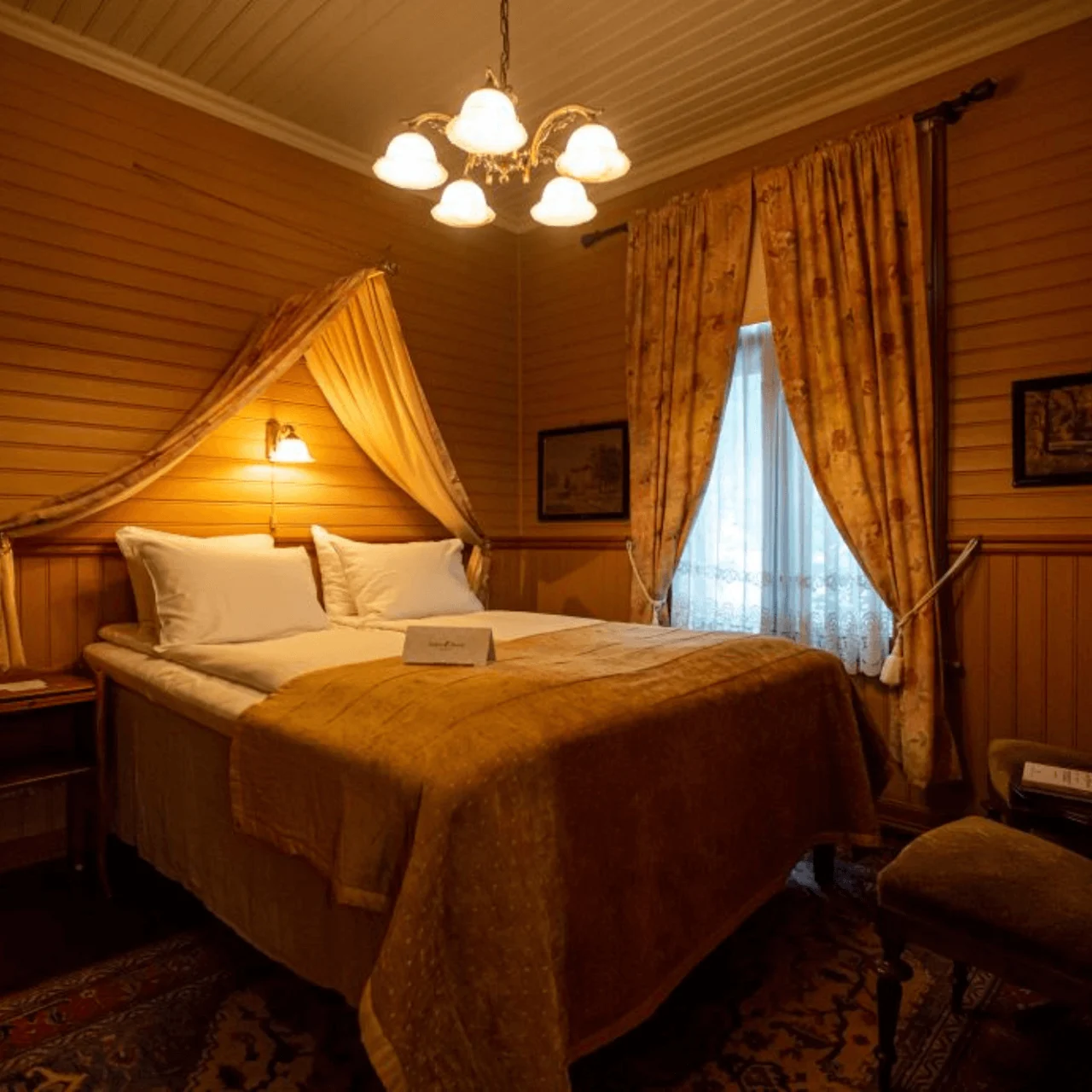 A large bed with white pillows and a yellow coverlet stands under a soft light in a warmly lit, vintage-style bedroom with wooden walls and patterned curtains.