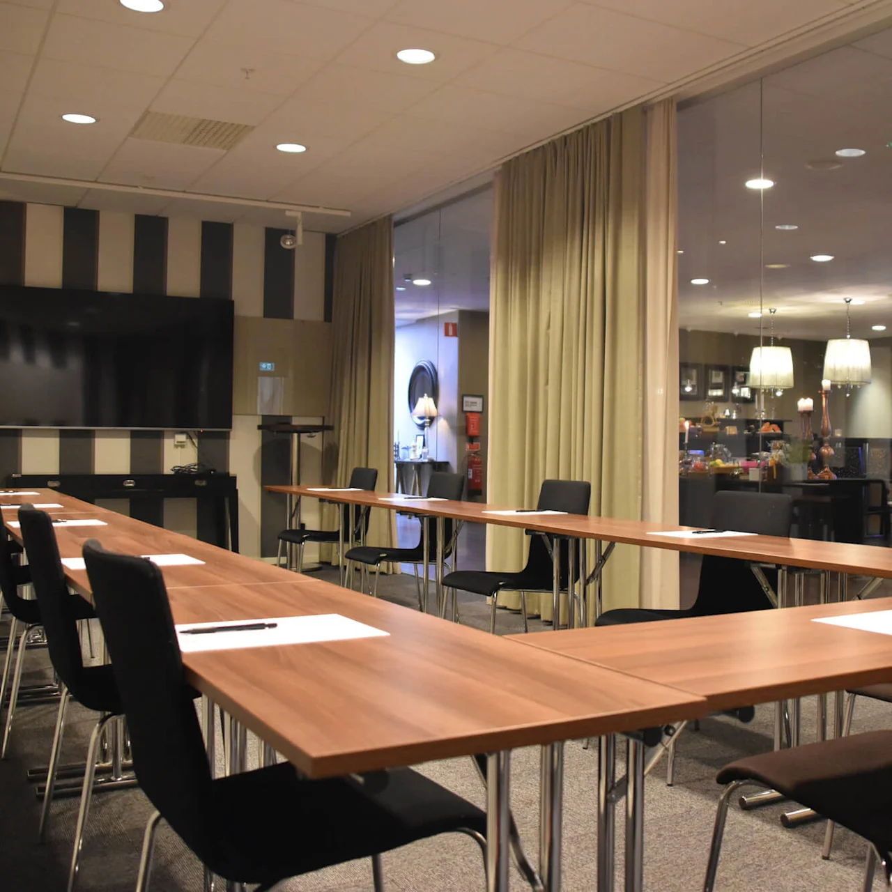 Konferencelokale med U-formede træborde, sorte stole, papir og penne, vendt mod en stor vægmonteret skærm. Omgivet af glasvægge, gardiner og synlige loftlamper. Ingen tekst synlig.