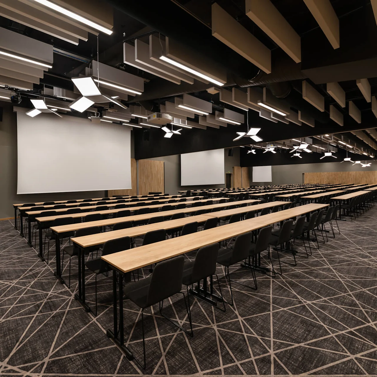 Rader av långa bord med svarta stolar är arrangerade i ett stort konferensrum. Rummet har flera skärmar och geometriska mattmönster, under en rad moderna taklampor.