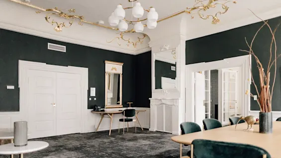 Elegant rum med mørke vægge, udsmykket guld- og hvidt loft, et stort bord med stole og en moderne lysekrone.
