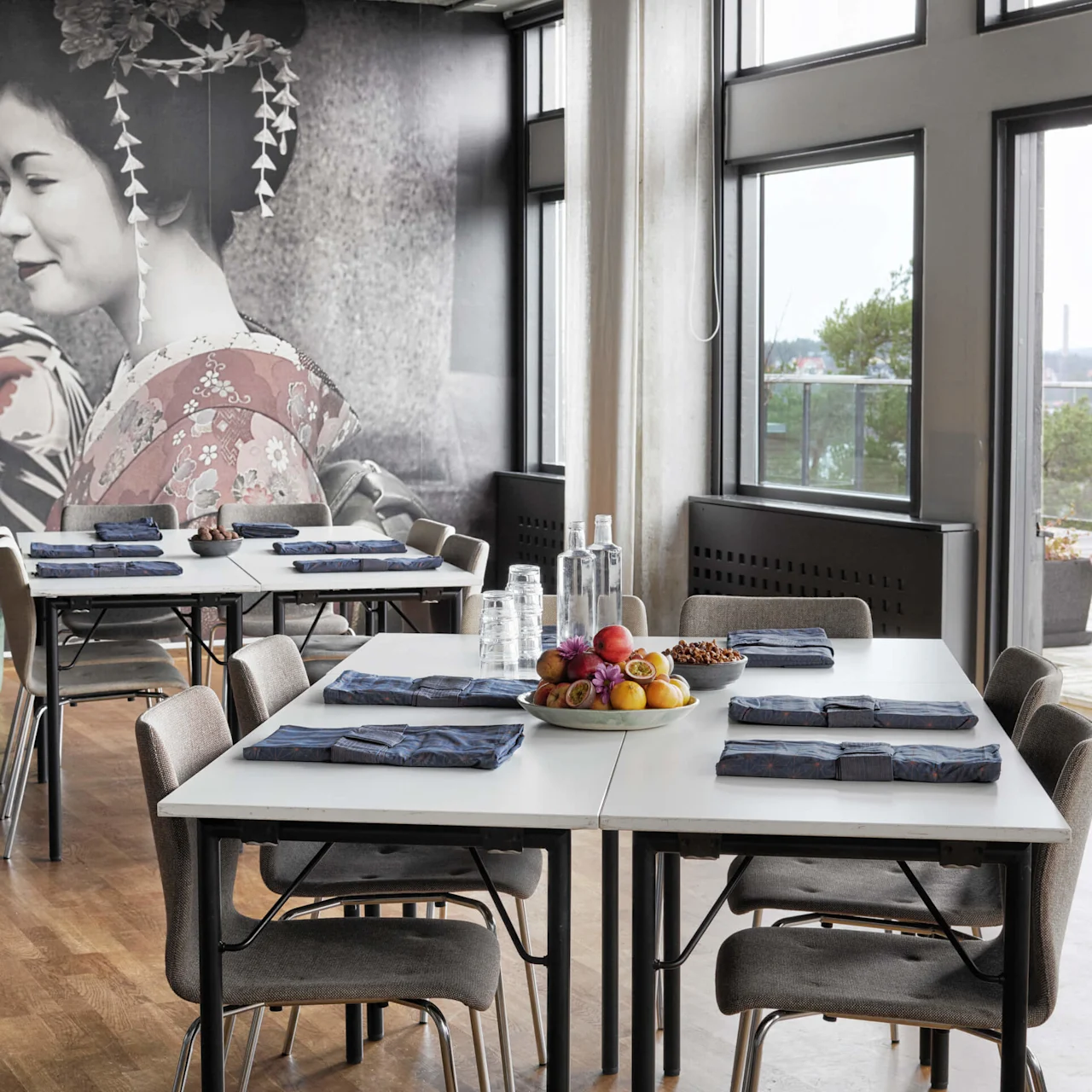 Et moderne spiseområde med dekkede bord, stoler rundt, og et stort veggkunstverk av geishaer. Store vinduer gir naturlig lys.