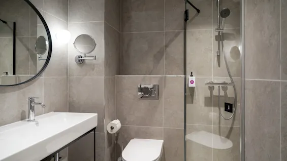 Et moderne bad med grå flislagte vegger, utstyrt med vask, toalett og en dusj med glassvegg.