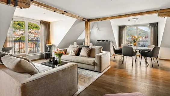 Et rummeligt opholdsområde har beige sofaer med puder, et mørkt sofabord med blomster og et spisebord ved store vinduer. Træbjælker pryder loftet med udsigt over en naturskøn udsigt.
