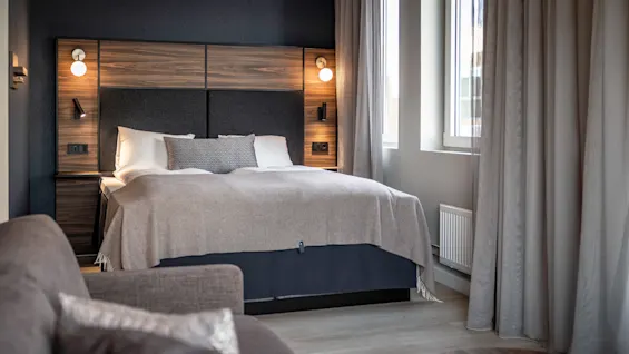 En säng med kuddar täckt av en beige filt står mot en mörk trärygg med inbyggda lampor. Den är belägen i ett modernt sovrum med stora fönster och gardiner.